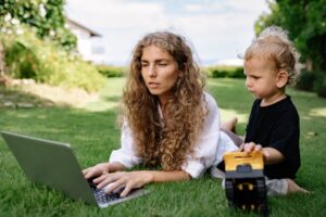 Woman working on a laptop while lying on grass with her child nearby playing.
